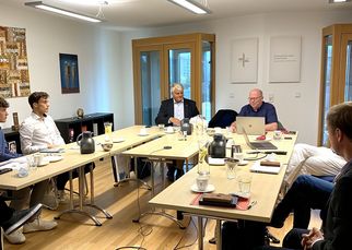 Eine Gruppe von sechs Personen sitzt an einem langen Tisch in einem Besprechungsraum. Einige haben Laptops und Notizen vor sich. Im Hintergrund sind Fenster und Kunstwerke an der Wand sichtbar. Die Atmosphäre wirkt konzentriert und professionell. (Text KI generiert)