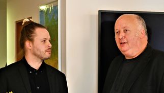 Zwei Männer in schwarzen Anzügen stehen in einem Raum und unterhalten sich. Der Mann links hat lange Haare, während der Mann rechts eine Glatze hat. Auf einem Tisch vor ihnen stehen Tulpen und andere Dekorationen. (Text KI generiert)