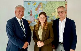 Drei Personen stehen vor einer Weltkarte. Zwei Männer in Anzügen, einer mit Krawatte, und eine Frau in einem braunen Anzug. Sie lächeln und posieren für das Foto. (Text KI generiert)