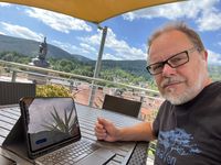 Ein Mann sitzt an einem Tisch im Freien, vor ihm ein Tablet mit Tastatur. Im Hintergrund sind Berge und eine Stadtlandschaft mit einem Kirchturm zu sehen. Der Himmel ist blau mit einigen Wolken. Der Mann zeigt mit dem Daumen nach oben. (Text KI generiert)