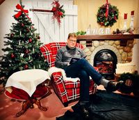Ein Mann sitzt in einem rot-weiß karierten Sessel vor einem Kamin. Er liest ein Buch, während ein Weihnachtsbaum und festliche Dekorationen im Hintergrund zu sehen sind. Der Raum ist gemütlich und weihnachtlich geschmückt. (Text KI generiert)