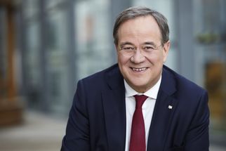 Ein lächelnder Mann in einem dunklen Anzug mit einer roten Krawatte sitzt vor einer modernen Glasarchitektur. Er hat kurze, graue Haare und trägt eine Brille. Der Hintergrund ist unscharf und zeigt einen urbanen Raum. (Text KI generiert)