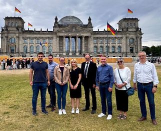 Eine Gruppe von sieben Personen steht auf einer Wiese vor dem Reichstagsgebäude in Berlin. Im Hintergrund sind deutsche Flaggen zu sehen. Die Personen sind lächelnd und in formeller Kleidung gekleidet. (Text KI generiert)
