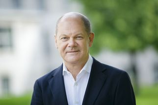 Ein Mann mit kurzen, grauen Haaren trägt einen dunklen Anzug und ein weißes Hemd. Er steht im Freien vor unscharfem, grünem Hintergrund und lächelt leicht. (Text KI generiert)
