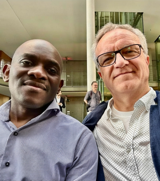 Zwei Männer posieren für ein Selfie in einem modernen Innenraum. Der Mann links trägt ein kariertes Hemd und hat einen ernsten Gesichtsausdruck, während der Mann rechts eine Brille trägt und lächelt. Im Hintergrund sind weitere Personen und große Fenster sichtbar.