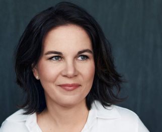 Porträt einer Frau mit dunklen, lockigen Haaren, die in einem weißen Hemd gekleidet ist. Sie sitzt an einem Tisch und schaut leicht lächelnd zur Seite. Der Hintergrund ist dunkel und neutral gehalten. (Text KI generiert)