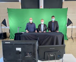 Drei Männer stehen hinter einem Tisch vor einem grünen Hintergrund. Sie tragen Masken und schauen in die Kamera. Vor ihnen stehen zwei Monitore. Die Umgebung wirkt wie ein Studio mit Beleuchtung. (Text KI generiert)
