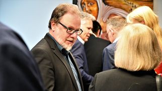 Eine Gruppe von Menschen steht in einem Raum und unterhält sich. Im Vordergrund ist ein Mann mit Brille und Bart zu sehen, der mit einer Person spricht. Im Hintergrund sind weitere Personen und ein Kunstwerk an der Wand sichtbar. (Text KI generiert)