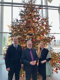 Drei Personen stehen vor einem festlich geschmückten Weihnachtsbaum in einem modernen Gebäude. Der Baum ist mit roten und goldenen Ornamenten sowie Lichtern dekoriert. Die Personen lächeln und tragen formelle Kleidung. (Text KI generiert)
