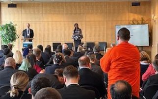 Eine Konferenzszene mit einem Publikum, das einem Vortrag zuhört. Auf der Bühne stehen zwei Personen, eine Frau spricht, während ein Mann daneben steht. Im Vordergrund ist ein Mann in orangefarbener Robe zu sehen, der sich von den anderen abhebt. (Text KI generiert)