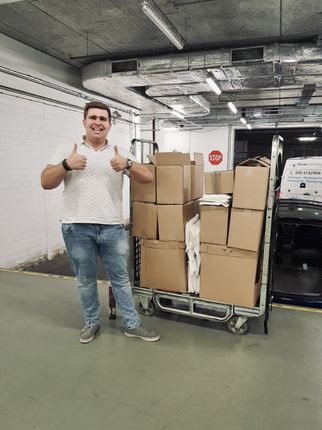 Ein lächelnder Mann steht neben einem Wagen voller Kartons in einer Parkgarage. Er zeigt mit beiden Daumen nach oben und wirkt zufrieden. Im Hintergrund ist ein Stoppschild sichtbar. (Text KI generiert)