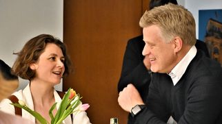 Zwei Personen unterhalten sich an einem Tisch. Eine Frau mit lockigem Haar lächelt und schaut den Mann an, der in einem schwarzen Pullover sitzt. Auf dem Tisch stehen eine Vase mit Tulpen, Snacks und Getränke. Im Hintergrund sind weitere Personen zu sehen. (Text KI generiert)
