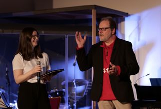 Eine Frau mit einem Mikrofon steht neben einem Mann, der gestikuliert. Beide lächeln und scheinen in ein Gespräch vertieft zu sein. Im Hintergrund sind Musikinstrumente und eine Bühne sichtbar. (Text KI generiert)