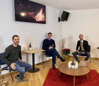 Drei Männer sitzen in einem modernen Raum. Im Hintergrund ist ein Bildschirm mit einem Feuer zu sehen. Auf einem Tisch stehen Kerzen und ein Laptop. Der Raum hat einen gemütlichen, festlichen Eindruck. (Text KI generiert)