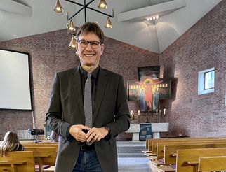 Ein Mann in einem schwarzen Anzug und Krawatte steht in einer Kirche mit Backsteinwänden. Im Hintergrund sind eine moderne Lichtinstallation und ein Altar mit religiösen Symbolen zu sehen. Bänke sind im Vordergrund sichtbar, einige Personen sitzen in der Kirche. (Text KI generiert)