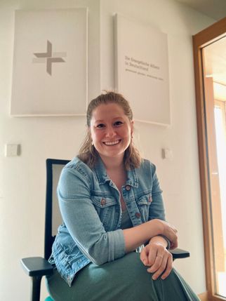 Eine lächelnde Frau sitzt auf einem Stuhl in einem hellen Raum. Sie trägt eine Jeansjacke und hat lange, lockige Haare. Im Hintergrund sind zwei Bilder an der Wand zu sehen, eines mit einem Kreuzsymbol und das andere mit Text. (Text KI generiert)