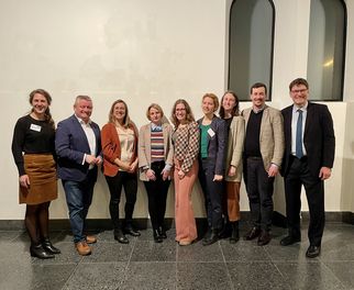 Eine Gruppe von neun Personen steht in einem Innenraum mit hellen Wänden. Sie lächeln und posieren für das Foto. Die Personen tragen verschiedene formelle und legere Kleidung. Im Hintergrund sind große Fenster zu sehen. (Text KI generiert)