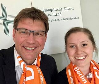 Zwei Personen lächeln in die Kamera, beide tragen orangefarbene Schals. Im Hintergrund ist ein Schild mit dem Text "Evangelische Allianz Deutschland" zu sehen. Die Umgebung wirkt freundlich und einladend. (Text KI generiert)