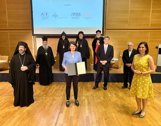 Eine Gruppe von Menschen steht auf einer Bühne. Im Vordergrund hält eine Frau ein Dokument in der Hand. Hinter ihr stehen mehrere Männer in religiöser Kleidung. Im Hintergrund ist eine Präsentationsleinwand mit Logos zu sehen. Die Szene scheint eine offizielle Veranstaltung zu sein. (Text KI generiert)