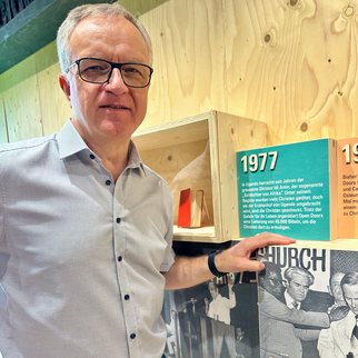 Ein Mann steht vor einer Wand mit Informationen zur Geschichte einer Kirche. Er zeigt auf ein Schild mit der Jahreszahl 1977 und erklärt die Bedeutung dieses Jahres für die Kirche und die Gemeinschaft. Im Hintergrund sind historische Fotos zu sehen.