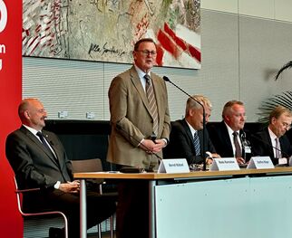 Eine Pressekonferenz mit mehreren Personen an einem Tisch. Ein Mann steht am Rednerpult und spricht, während die anderen vier Männer ihm zuhören. Im Hintergrund ist ein großes Kunstwerk zu sehen. An der Wand ist ein rotes Banner mit dem Schriftzug "SPD".