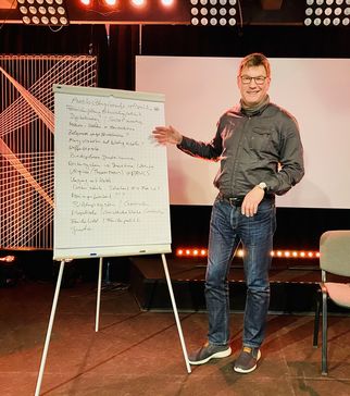 Ein Mann steht neben einem Flipchart, auf dem handschriftliche Notizen stehen. Er trägt eine graue Jacke und Jeans. Im Hintergrund sind Lichter und eine leere Stuhlreihe zu sehen. Die Szene scheint in einem Veranstaltungsraum zu sein. (Text KI generiert)