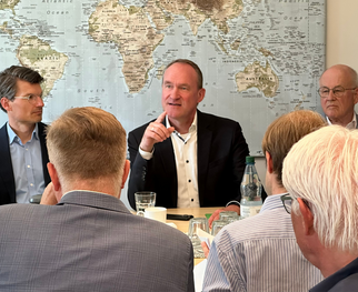 Eine Gruppe von Männern sitzt an einem Tisch, während einer von ihnen spricht. Im Hintergrund ist eine Weltkarte zu sehen. Die Männer tragen formelle Kleidung und scheinen an einer Diskussion oder einem Meeting teilzunehmen.