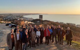 Eine Gruppe von etwa 30 Personen steht auf einem Hügel mit Blick auf eine Küstenstadt bei Sonnenuntergang. Die Menschen lächeln und posieren für das Foto, während im Hintergrund das Meer und die Stadt sichtbar sind. (Text KI generiert)