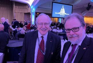 Zwei Männer posieren für ein Selfie in einem festlich dekorierten Raum mit Tischen, die für ein Event vorbereitet sind. Im Hintergrund ist eine große Leinwand mit dem Schriftzug "THE NATIONAL PRESS BREAKFAST" und einer Abbildung des Kapitols zu sehen. (Text KI generiert)