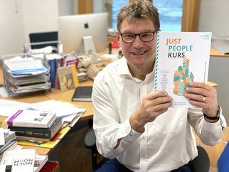 Ein Mann mit Brille sitzt an einem Schreibtisch und hält ein Buch mit dem Titel "JUST PEOPLE KURS" in der Hand. Der Schreibtisch ist mit verschiedenen Unterlagen und Büchern bedeckt. Im Hintergrund ist ein Computer sichtbar. (Text KI generiert)