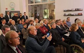 Eine Gruppe von Menschen sitzt in einem Raum, um an einer Veranstaltung teilzunehmen. Die Anwesenden sind unterschiedlich alt und tragen formelle Kleidung. Einige schauen nach vorne, während andere mit ihren Handys beschäftigt sind. Im Hintergrund sind weitere Personen zu sehen. (Text KI generiert)