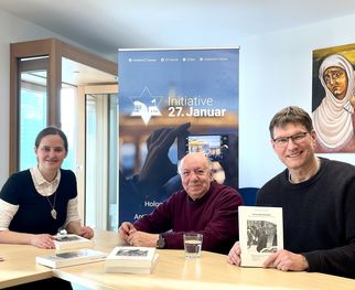 Drei Personen sitzen an einem Tisch, umgeben von Büchern. Im Hintergrund steht ein Banner mit der Aufschrift "Initiative 27. Januar". Eine Frau in einem schwarzen und weißen Outfit lächelt, während ein älterer Mann in einem roten Pullover und ein Mann in einem grauen Pullover Bücher halten. (Text KI generiert)