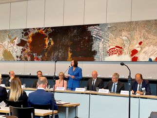 Eine Frau in blauer Kleidung steht an einem Rednerpult vor einer Gruppe von Menschen, die an einem Tisch sitzen. Im Hintergrund ist ein großes, abstraktes Wandbild zu sehen. Die Anwesenden scheinen an einer Diskussion oder Präsentation teilzunehmen. (Text KI generiert)