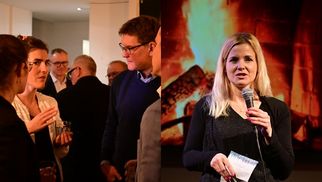 Eine Gruppe von Menschen unterhält sich in einem Raum, während eine Frau mit Mikrofon auf einer Bühne spricht. Im Hintergrund ist ein Kaminfeuer zu sehen. (Text KI generiert)