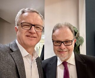 Zwei Männer stehen nebeneinander und lächeln in die Kamera. Der Mann links trägt ein graues Sakko und ein helles Hemd, der Mann rechts hat einen Anzug mit einer lila Krawatte an. Im Hintergrund sind unscharfe Elemente eines Innenraums zu sehen. (Text KI generiert)