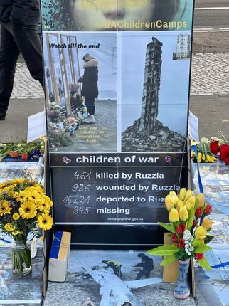 Eine Gedenkstätte mit Bildern und Blumen, die auf die Auswirkungen des Krieges auf Kinder hinweist. Ein Plakat zeigt die Zahlen: 461 getötet, 926 verwundet, 16.221 deportiert und 345 vermisst. Der Slogan "Watch till the end" ist ebenfalls sichtbar. (Text KI generiert)