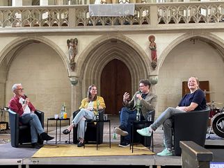 Vier Personen sitzen in einem historischen Raum auf schwarzen Stühlen. Sie diskutieren und lachen, während ein Mikrofon auf einem Tisch vor ihnen steht. Im Hintergrund sind architektonische Details und eine Musikanlage sichtbar. (Text KI generiert)