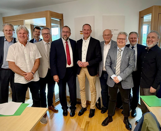 Eine Gruppe von zehn Männern steht in einem Raum mit großen Fenstern. Sie tragen formelle Kleidung und lächeln in die Kamera. Auf einem Tisch im Vordergrund liegen einige Dokumente und grüne Ordner.