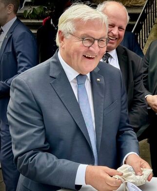 Ein lächelnder Mann in einem grauen Anzug mit Brille hält eine kleine Tasche in der Hand. Im Hintergrund stehen weitere Personen, die ebenfalls lächeln. Die Szene wirkt freundlich und feierlich. (Text KI generiert)