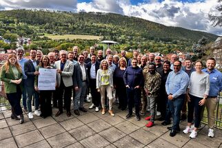 Eine große Gruppe von Menschen steht auf einer Terrasse mit Blick auf eine grüne Landschaft und Hügel. Einige Personen halten ein Dokument in der Mitte der Gruppe. Es ist sonnig und die Wolken sind teilweise bewölkt. (Text KI generiert)
