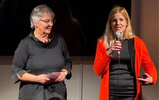 Zwei Frauen stehen auf einer Bühne. Eine Frau trägt ein schwarzes Kleid und hält ein Blatt Papier, während die andere in einem roten Oberteil mit Mikrofon spricht. Im Hintergrund ist ein dunkler Bildschirm sichtbar. (Text KI generiert)