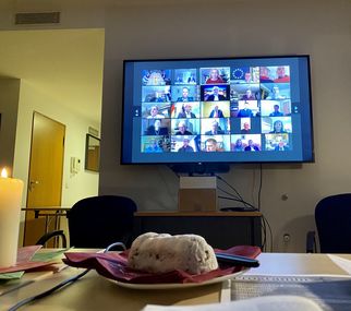 Ein Tisch mit einem roten Teller, auf dem ein Stück Kuchen liegt. Im Hintergrund ist ein Bildschirm zu sehen, der eine Videokonferenz mit mehreren Teilnehmern zeigt. Eine Kerze brennt auf dem Tisch. (Text KI generiert)