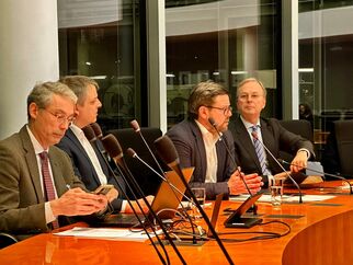Eine Gruppe von vier Männern sitzt an einem Tisch in einem Konferenzraum. Zwei von ihnen sprechen, während die anderen zuhören. Auf dem Tisch befinden sich Mikrofone, Laptops und Gläser mit Wasser. Große Fenster im Hintergrund lassen Tageslicht herein. (Text KI generiert)