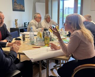 Eine Gruppe von fünf Personen sitzt an einem Tisch in einem modernen Büro. Sie diskutieren und haben Getränke sowie Unterlagen vor sich. Im Hintergrund sind Fenster mit Blick auf eine Stadtlandschaft.