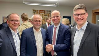 Vier Männer stehen in einem modernen Raum, lächeln und posieren für ein Foto. Im Hintergrund sind weitere Personen und eine dekorative Wand zu sehen. Die Männer tragen formelle Kleidung und scheinen an einem gesellschaftlichen Ereignis teilzunehmen. (Text KI generiert)