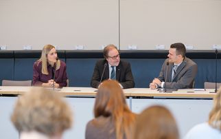 Drei Personen sitzen an einem Tisch in einem Konferenzraum. Zwei Männer und eine Frau diskutieren, während eine Gruppe von Zuhörern im Hintergrund sitzt. Die Frau trägt ein lila Outfit, der Mann in der Mitte hat eine Brille und einen Anzug, der dritte Mann trägt einen grauen Anzug. (Text KI generiert)