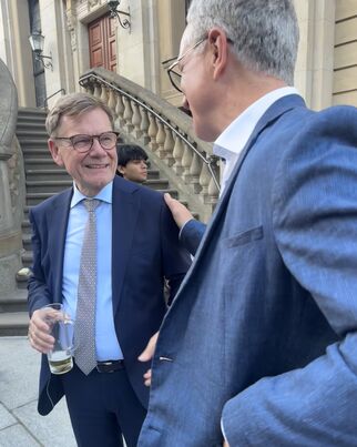 Zwei Männer stehen an einer Treppe und unterhalten sich. Der Mann links trägt einen Anzug und hält ein Glas in der Hand. Der Mann rechts trägt ebenfalls einen Anzug und lächelt. Im Hintergrund ist eine Person zu sehen.