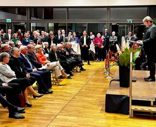 Eine Person steht auf einer Bühne und spricht zu einem Publikum in einem großen Raum. Die Zuhörer sitzen in Reihen und schauen aufmerksam zu. Im Hintergrund sind Pflanzen und Dekorationen sichtbar. Die Atmosphäre wirkt feierlich und formal. (Text KI generiert)