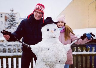 Ein Mann und ein Mädchen stehen neben einem Schneemann im Schnee. Der Mann trägt eine rote Mütze und einen schwarzen Mantel, während das Mädchen eine rosa Jacke und eine Mütze trägt. Der Schneemann hat einen schwarzen Hut und eine Karottennase. (Text KI generiert)
