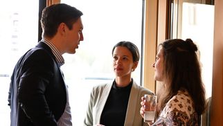 Drei Personen stehen in einem Raum und unterhalten sich. Ein Mann in einem Anzug spricht mit zwei Frauen, eine trägt ein helles Oberteil und die andere ein gemustertes Hemd. Eine der Frauen hält ein Glas Wasser in der Hand. (Text KI generiert)